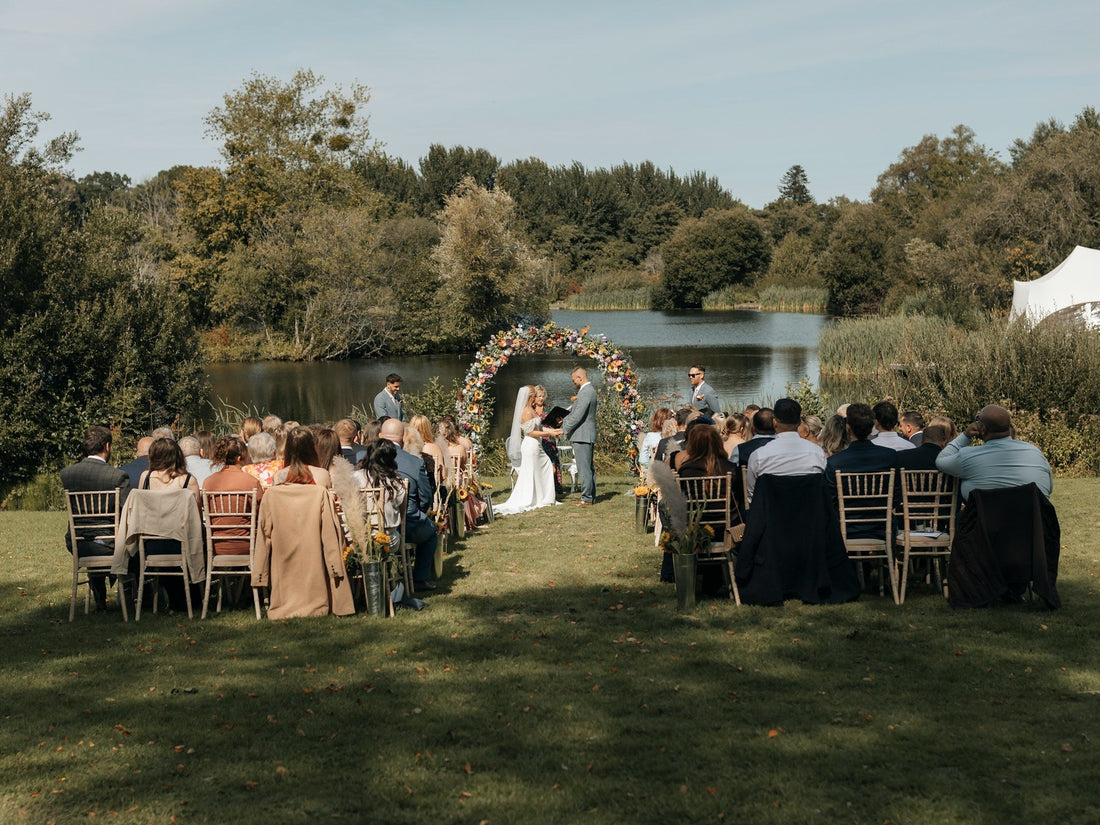 Beautiful outdoor wedding ceremony by a lake with guests seated, featuring the groom and groomsmen in tailored blue wedding suits.