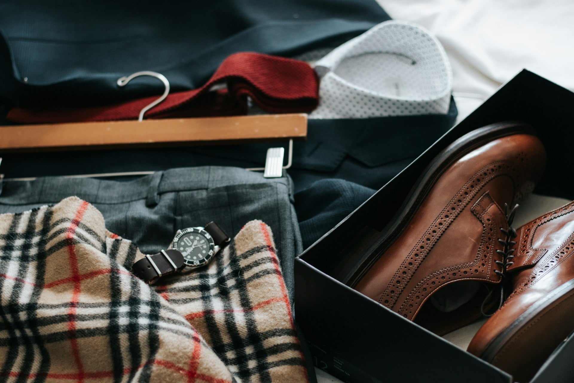 A flat lay of men's formalwear ready to be packed, featuring a dark suit, red knit tie, grey trousers, watch, and brown leather shoes, illustrating how to pack a suit for travel.