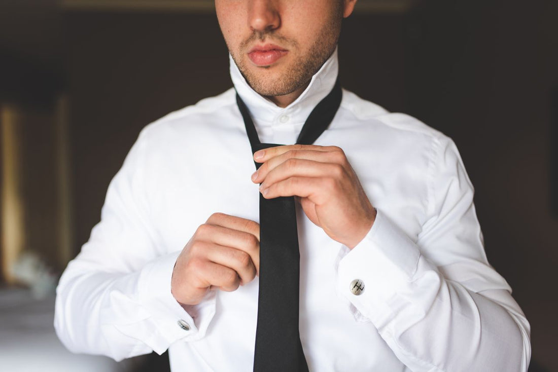 A close-up of a man in a crisp white dress shirt with French cuffs adjusting the knot of a black tie, demonstrating how to perfectly tie a tie for a suit.