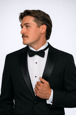 Men's black slim fit tuxedo featuring a classic two-button front with satin notch lapels and side vents for refined movement. The flat-front trousers showcase a satin side stripe and expandable waistband for comfort throughout the evening. An included dress shirt ensures you're completely outfitted for any black-tie occasion.