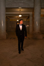 Men's black slim fit tuxedo featuring a classic two-button front with satin notch lapels and side vents for refined movement. The flat-front trousers showcase a satin side stripe and expandable waistband for comfort throughout the evening. An included dress shirt ensures you're completely outfitted for any black-tie occasion.