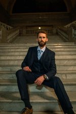 Man modeling a Men's Navy Tweed Slim Fit Suit with vest and floral tie on marble steps, formalwear style